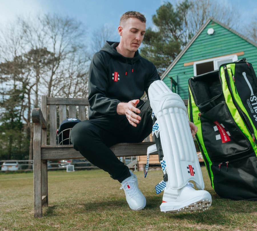 Gray-Nicolls campaign cricketer putting on batting pads beside a kit bag